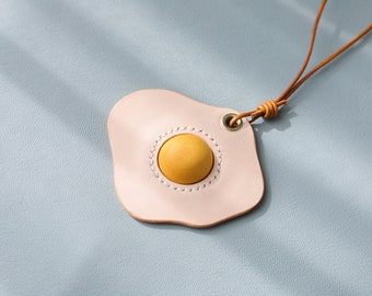 May include: A light pink leather keychain shaped like a fried egg with a yellow leather yolk. The keychain is attached to a brown leather cord.