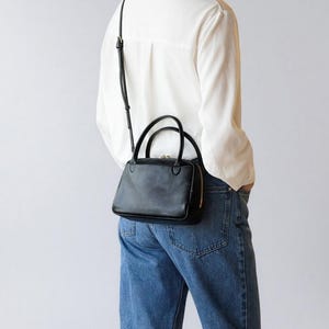 Könnte beinhalten: Eine schwarze Lederhandtasche mit einem oberen Griff und einem abnehmbaren Schulterriemen. Die Tasche hat eine rechteckige Form mit einem goldenen Reißverschluss und wird über der Schulter getragen. Die Person trägt ein weißes Hemd und blaue Jeans.