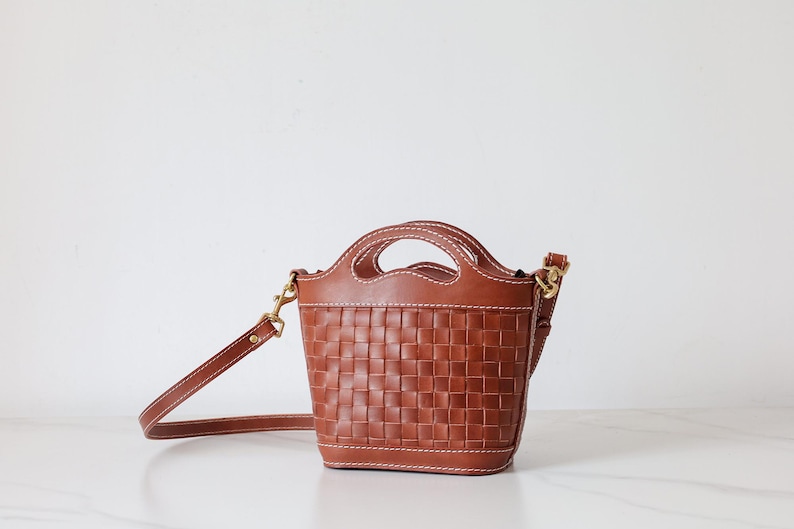 Puede incluir: Un bolso de mano de cuero marr&oacute;n con un dise&ntilde;o de cesta tejida. El bolso tiene un asa superior y una correa para el hombro desmontable. Las costuras son blancas y los herrajes son dorados. El bolso est&aacute; sobre una superficie blanca.
