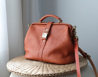 May include: A brown leather doctor bag with a gold buckle and a long strap. The bag is sitting on a woven straw surface.