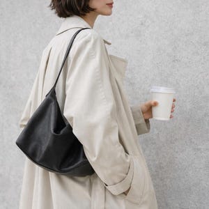 May include: A black leather shoulder bag with a curved design is worn over the shoulder. The person is wearing a light beige trench coat and holding a white paper coffee cup. The background is a light gray textured wall.