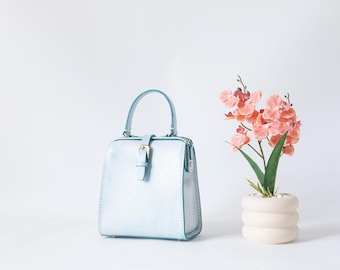 May include: A light blue handbag with a top handle and a buckle closure. The bag is next to a white vase with pink flowers and green leaves. The bag has a rectangular shape.