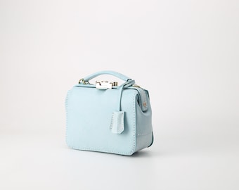 May include: A light blue leather handbag with a top handle and a small, decorative flap. The bag has a silver clasp and a gold zipper.
