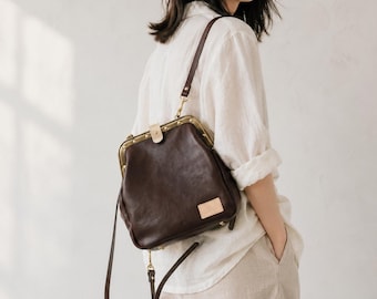 May include: A dark brown leather satchel bag with a clasp closure and a long, adjustable strap. The bag has a rectangular shape with rounded edges and a small, rectangular tag on the front. The hardware is gold-colored.