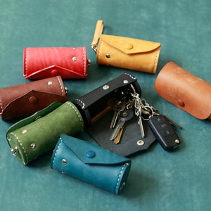 May include: An assortment of colourful leather keychains in various colours, including red, brown, green, blue, and yellow. The keychains are cylindrical in shape and have a flap closure. One keychain is open and has a set of keys and a car key fob attached.