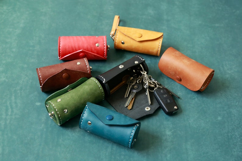 May include: An assortment of colourful leather keychains in various colours, including red, brown, green, blue, and yellow. The keychains are cylindrical in shape and have a flap closure. One keychain is open and has a set of keys and a car key fob attached.
