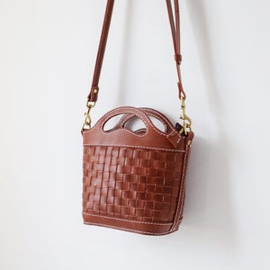 Puede incluir: Bolso de mano de cuero marr&oacute;n con dise&ntilde;o tejido. El bolso tiene un asa superior y una correa para el hombro desmontable. Mide aproximadamente 20 cm de alto y 18 cm de ancho. Cuelga sobre un fondo blanco.