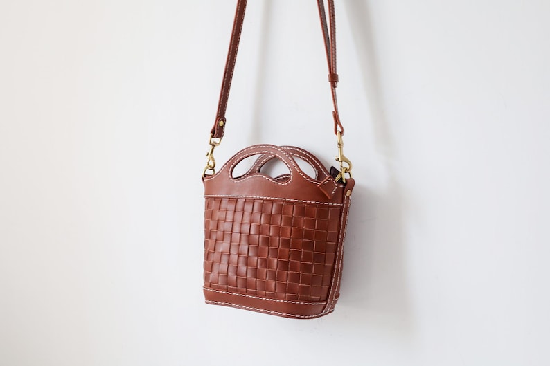 Puede incluir: Bolso de mano de cuero marr&oacute;n con dise&ntilde;o tejido. El bolso tiene un asa superior y una correa para el hombro desmontable. Mide aproximadamente 20 cm de alto y 18 cm de ancho. Cuelga sobre un fondo blanco.