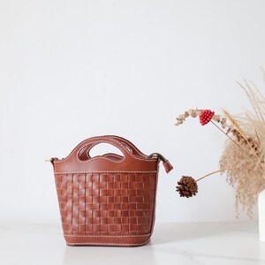 Puede incluir: Un bolso de mano de cuero marr&oacute;n con un dise&ntilde;o tejido. El bolso tiene dos asas y una correa para el hombro. Est&aacute; sobre una superficie blanca. Un jarr&oacute;n blanco con flores secas y una pi&ntilde;a se encuentra a la derecha del bolso.