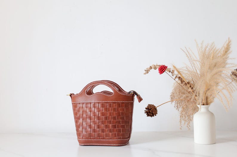 Puede incluir: Un bolso de mano de cuero marr&oacute;n con un dise&ntilde;o tejido. El bolso tiene dos asas y una correa para el hombro. Est&aacute; sobre una superficie blanca. Un jarr&oacute;n blanco con flores secas y una pi&ntilde;a se encuentra a la derecha del bolso.