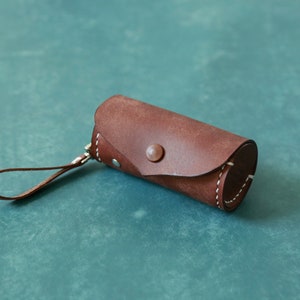 May include: A brown leather cylindrical case with white stitching detail and a silver button closure. The case has a brown leather strap.