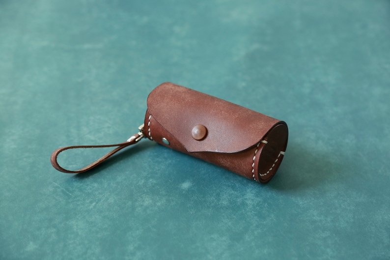 May include: A brown leather cylindrical case with white stitching detail and a silver button closure. The case has a brown leather strap.