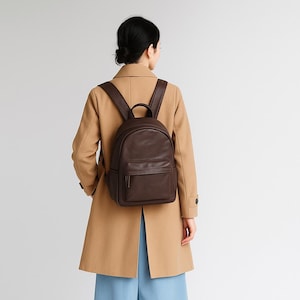 May include: A brown leather backpack with a front pocket and adjustable straps. The backpack is worn by a person wearing a tan coat and blue pants. The bag has a top handle and a zipper closure on the front pocket.