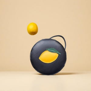 May include: A navy blue round handbag with a lemon applique. The lemon is yellow with a green leaf. A whole lemon floats above the bag. The bag has a handle and white stitching.