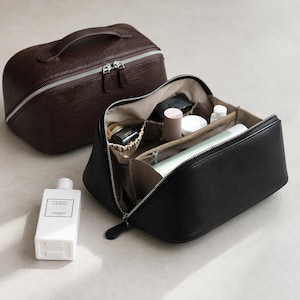 May include: A dark brown leather travel bag with a top handle and silver zip, and an open black leather travel bag containing various small bottles and containers, alongside a white rectangular bottle labelled Un Jardin sur le Nil Hermes.
