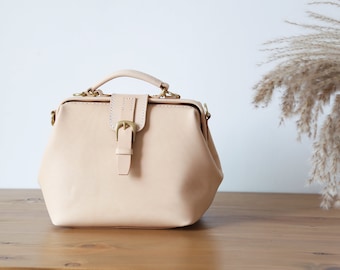 May include: A light beige leather doctor bag with a brass buckle and a top handle. The bag is sitting on a wooden surface.