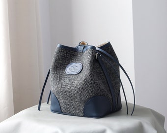 May include: A gray and blue bucket bag with a herringbone pattern and leather trim. The bag has a drawstring closure and two long straps.