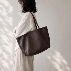 May include: A large, dark brown leather tote bag with two thin shoulder straps. The bag features a simple, open-top design with a smooth, matte finish and a rectangular shape, hanging from the shoulder with a spacious main compartment.
