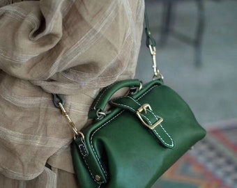 May include: A dark green leather doctor bag with a gold buckle and strap. The bag has white stitching around the edges.
