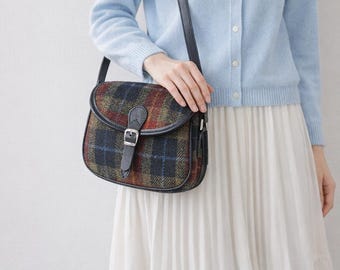 May include: A shoulder bag with a plaid pattern in shades of red, green, and navy blue. The bag has a black leather strap and buckle closure. The bag is worn over a light blue cardigan.