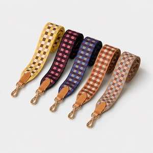 May include: Five adjustable bag straps featuring a checkered pattern in yellow, black and pink, blue and black, orange and brown, and light blue and tan. Each strap includes tan leather tabs and gold-toned metal swivel lobster claw clasps.