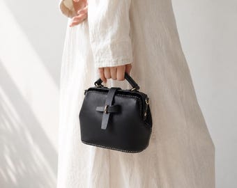 May include: A black structured handbag with a top handle and a single strap closure featuring a brass-toned metal ring. The bag has visible white stitching along the bottom edge and sides, with a boxy silhouette and a secure frame opening.