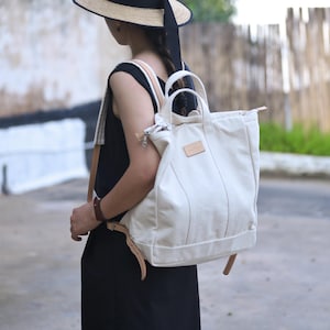 May include: A white canvas tote bag with brown leather straps and a brown leather handle. The bag is worn as a backpack.