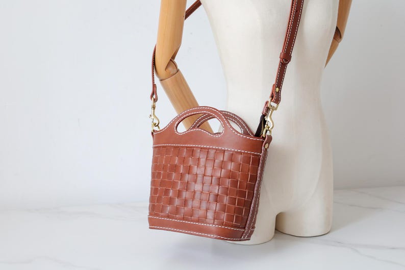 Puede incluir: Bolso bandolera de cuero marr&oacute;n con dise&ntilde;o tejido. El bolso tiene un asa superior y una correa para el hombro desmontable. El bolso mide aproximadamente 20 cm de alto y 18 cm de ancho. El bolso se muestra en un maniqu&iacute;.