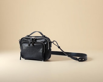 May include: A black leather crossbody bag with a top handle and a long, adjustable strap. The bag has a rectangular shape with a zippered main compartment and an exterior pocket. The hardware is silver-toned.