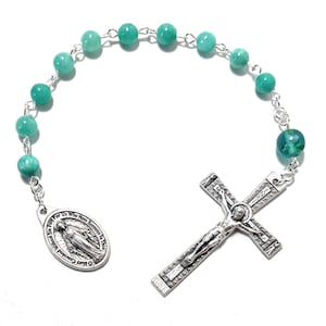 May include: A rosary with turquoise-colored beads and a silver crucifix. The crucifix features a detailed depiction of Jesus Christ. An oval pendant with text and an image of the Virgin Mary is also present.