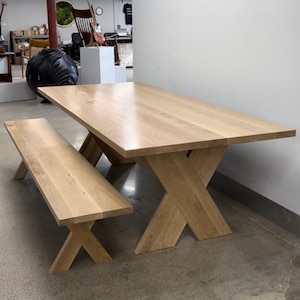 Puede incluir: Un juego de mesa y banco de comedor de madera clara con una base en forma de X. La mesa y el banco están hechos de madera maciza y tienen un acabado natural.
