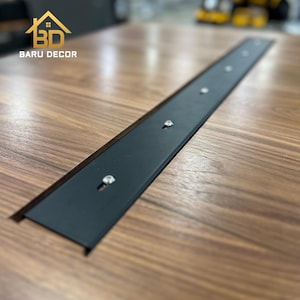 May include: A close-up shot of a wooden table with a black metal strip inlaid. The metal strip is secured with silver screws. The table has a rich, brown wood grain pattern. The logo "BARU DECOR" is in the top left corner.