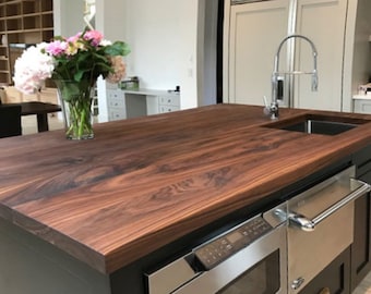 May include: A close-up of a dark brown walnut kitchen island countertop with a stainless steel sink and faucet. The countertop has a natural wood grain pattern and is surrounded by black cabinets.