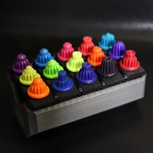 May include: A collection of colorful plastic caps arranged in a grid on a gray platform. The caps are in various shades of red, orange, yellow, green, blue, and purple.