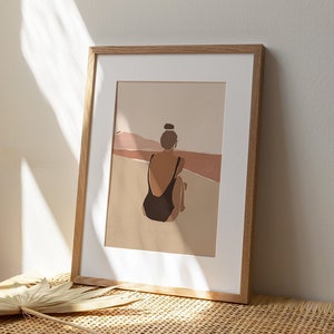 May include: A framed illustration of a person in a black swimsuit sitting on a beach, looking out at a landscape of brown and pink hills.