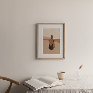May include: A framed illustration of a person in a black swimsuit sitting on a beach, facing the ocean. The background is a muted orange and pink color.