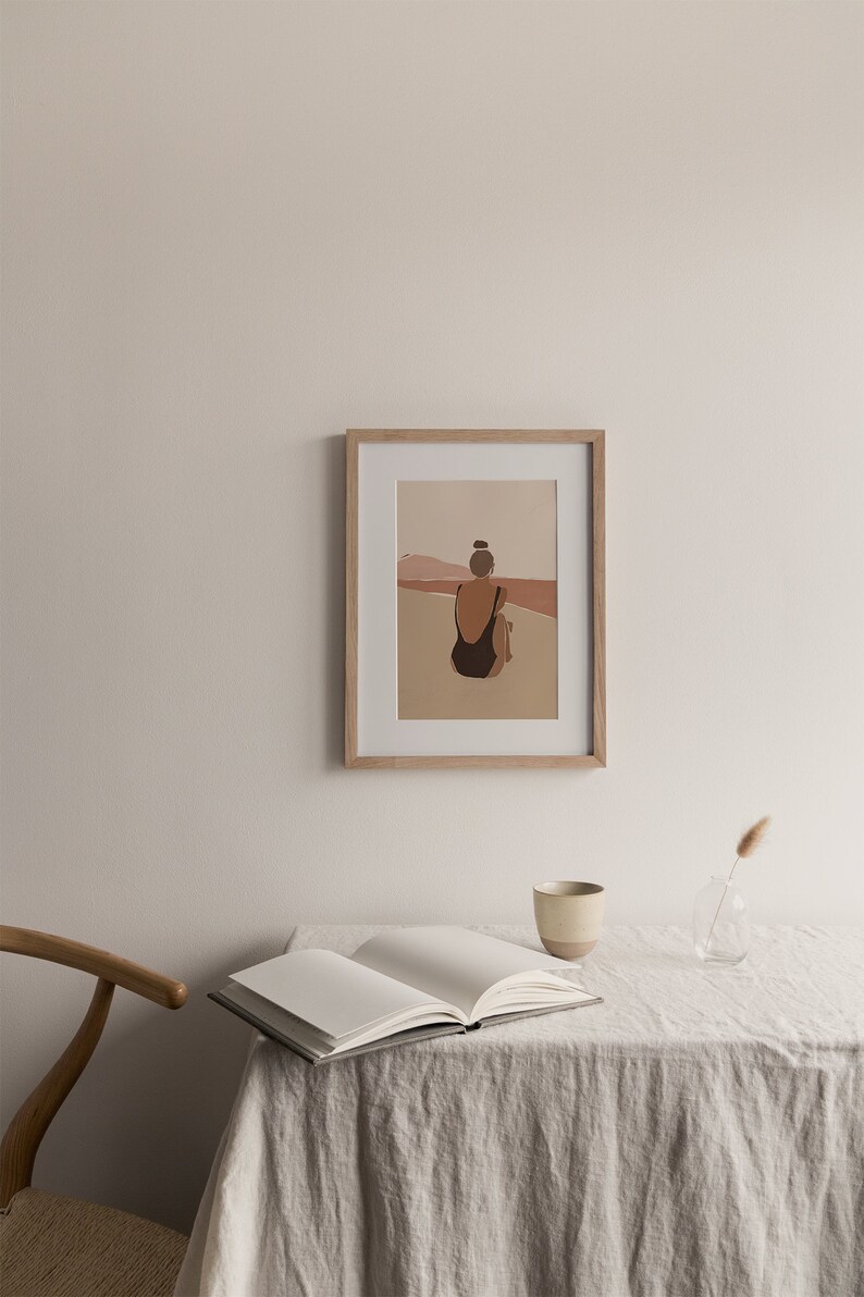 May include: A framed illustration of a person in a black swimsuit sitting on a beach, facing the ocean. The background is a muted orange and pink color.