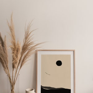 May include: A framed minimalist abstract print with a black circle and a thin black line on a beige background. The print is set against a white wall with a vase of dried pampas grass in front of it.