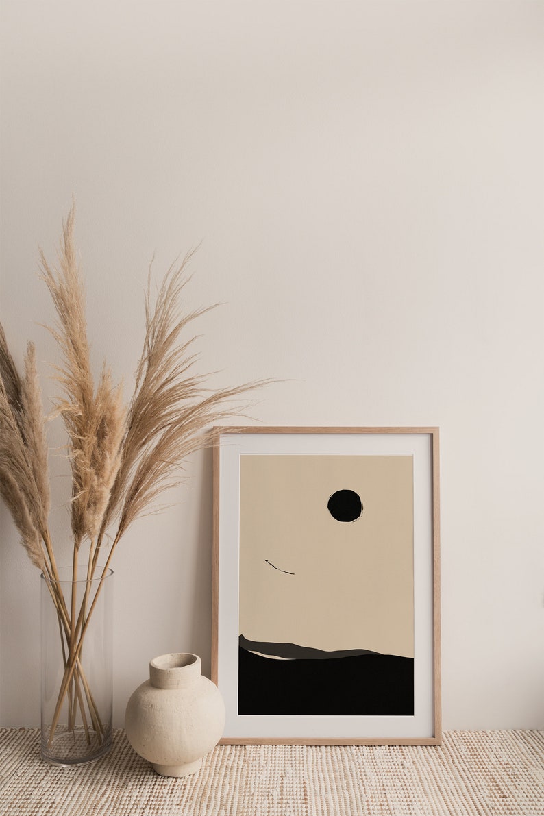 May include: A framed minimalist abstract print with a black circle and a thin black line on a beige background. The print is set against a white wall with a vase of dried pampas grass in front of it.