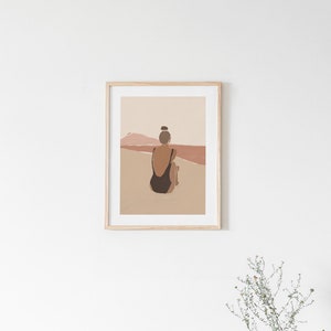 May include: A framed illustration of a person in a black swimsuit sitting on a beach, looking out at a sunset with a pink and orange sky.