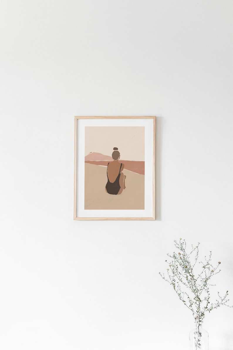 May include: A framed illustration of a person in a black swimsuit sitting on a beach, looking out at a sunset with a pink and orange sky.