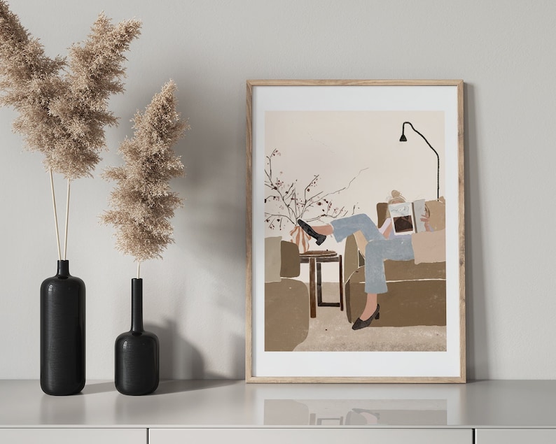 May include: A framed illustration of a person relaxing on a couch, reading a book. The illustration is in a minimalist style with muted colors and simple lines. The person is wearing blue jeans and a white shirt. The couch is brown and the background is a light beige.