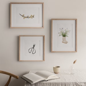 K&ouml;nnte beinhalten: Drei gerahmte Aquarell-Illustrationen an einer wei&szlig;en Wand. Die Illustrationen zeigen einen Zweig mit gelben Bl&auml;ttern, einen Blumenstrau&szlig; in einem wei&szlig;en Gummistiefel und eine schwarze Schere.