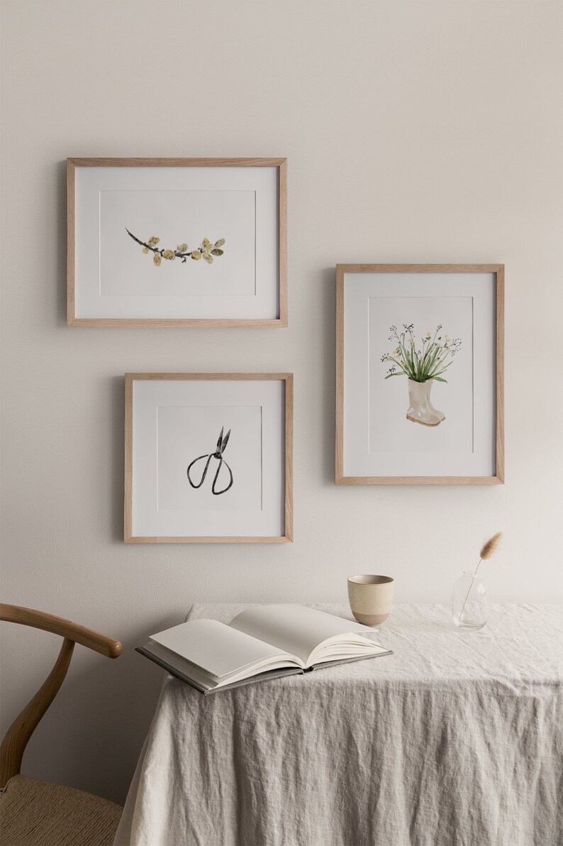 K&ouml;nnte beinhalten: Drei gerahmte Aquarell-Illustrationen an einer wei&szlig;en Wand. Die Illustrationen zeigen einen Zweig mit gelben Bl&auml;ttern, einen Blumenstrau&szlig; in einem wei&szlig;en Gummistiefel und eine schwarze Schere.