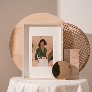 May include: A framed illustration of a woman in a green shirt and white pants standing in front of a leafy background. The frame is on a white table with a wooden tray and a round gold mirror.