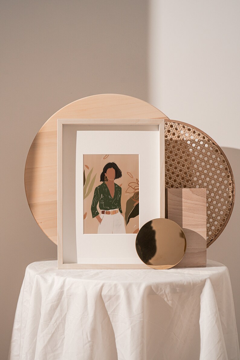 May include: A framed illustration of a woman in a green shirt and white pants standing in front of a leafy background. The frame is on a white table with a wooden tray and a round gold mirror.