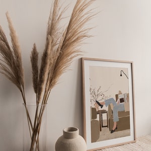 May include: A framed illustration of a person sitting on a couch reading a book. The illustration is in a minimalist style with muted colors. The frame is light wood and the illustration is on a beige background.