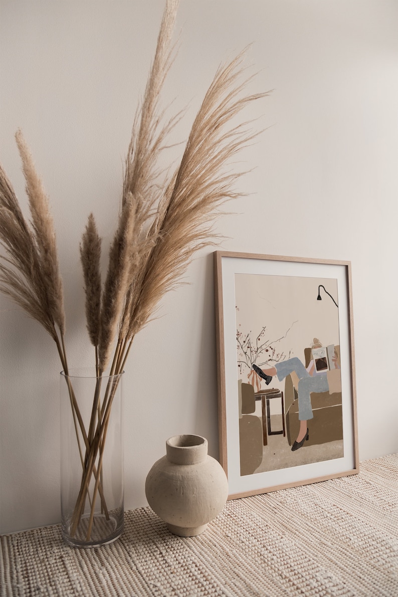 May include: A framed illustration of a person sitting on a couch reading a book. The illustration is in a minimalist style with muted colors. The frame is light wood and the illustration is on a beige background.