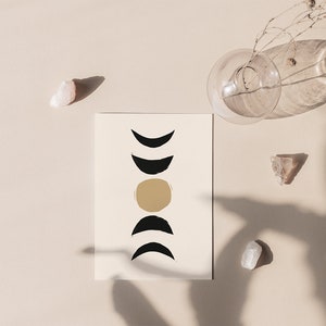 May include: A beige art print featuring a minimalist design of moon phases in black and gold. The print is accompanied by clear crystals and a glass vase with dried stems, all arranged on a neutral-toned surface.
