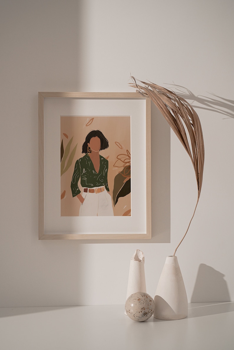 May include: A framed illustration of a woman wearing a green shirt and white pants standing in front of a floral background. The frame is on a white surface with two white ceramic vases and a decorative stone.